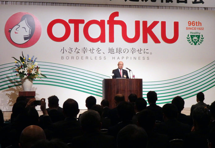新ロゴの前で方針を語る佐々木直義社長（オタフクソース）