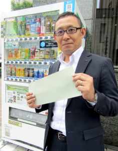 茶殻抗菌シールを持つ自販機部の杉浦義典副部長(伊藤園)。1枚で自販機1台分に対応。購入ボタン・おつり返却レバー・紙幣投入口・釣り銭口・商品取り出し口の各貼付箇所向けに切り抜きが施されている。