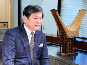 川股一雄副社長(キーコーヒー)。右はトラジャ地方に伝わる船形屋根の高床式家屋「トンコナン」の模型。座る場所を意味するトンコナンは集会所として利用される