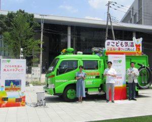 えきまえテラスに設置される「こども気温」を知らせる緑色の消防車「熱中症防車」。右から八王子市の初宿和夫（しやけ かずお）市長、サントリー食品インターナショナルの阿部泰丈ブランドマーケティング本部部長、多胡氏 - 食品新聞 WEB版（食品新聞社）