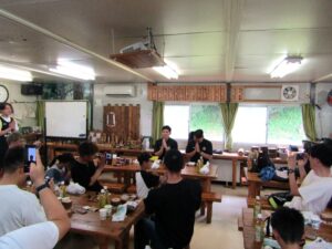 お昼にいただきますの挨拶をする細川一輝選手左奥と池田親平アシスタントコーチ兼スキルコーチ右奥 お昼に「いただきます」の挨拶をする細川一輝選手(左奥)と池田親平アシスタントコーチ兼スキルコーチ(右奥)