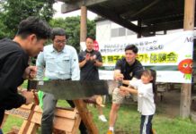 味の素AGF、「AGFブレンディの森」が広がる東西の生産拠点で地域貢献活動を深化 丸太切り体験の様子。子どもをサポートする細川一輝選手(右から2番目)見守る池田親平アシスタントコーチ兼スキルコーチ(中央奥)、丸太に乗って丸太を安定させる赤城南麓森林組合の石塚征人理事兼事業部部長