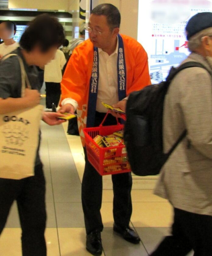 10日、東京駅一番街地下1階ののぞみ広場で道行く人に「亀田の柿の種 うましお」の試食品を手渡す亀田製菓の髙木社長COO