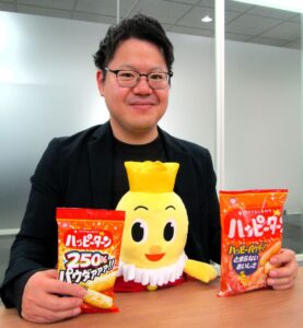 机の間に挟んでデスクワークすると姿勢がよくなりハッピー！になれるという「背筋ピーン！ぬいぐるみ ターン王子」を実演する歴舎（れきしゃ）直輝マーケティング戦略部米菓スナック第2グループマネージャー