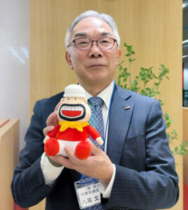R-1の新キャラクター「アールおじさん」を手に笑顔を見せる八尾文二郎社長