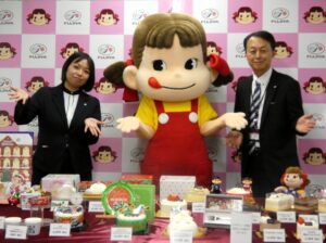 9月29日、不二家本社で開催された「2026年度 不二家洋菓子店クリスマスケーキお披露目会」に臨んだ金田剛執行役員洋菓子事業本部営業本部長（右）と瓜生麻未洋菓子事業本部営業本部マーケティング部営業推進課課長（左）。「ペコちゃん」を囲む。